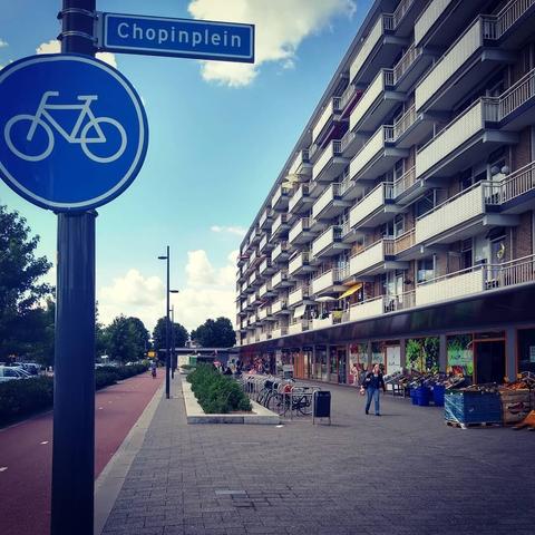 Rechts een flat met zes woonlagen boven kleurrijke winkeltjes. Op de voorgrond een straatnaambord: 'Chopinplein'