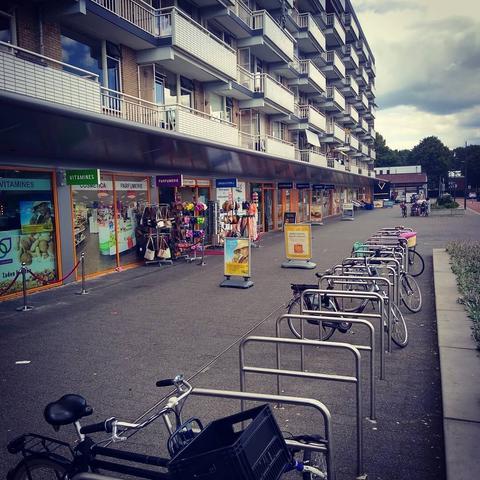 Links een flat met zes woonlagen boven kleurrijke winkeltjes. Er hangen gekleurde bordjes die uit de gevel steken bij de winkels: 'Vitamines', 'Parfumerie', 'Drogisterij' en de bordjes verder naar achteren zijn niet te lezen. Op de voorgrond beugels waar wat fietsen tegenaan staan gestald.