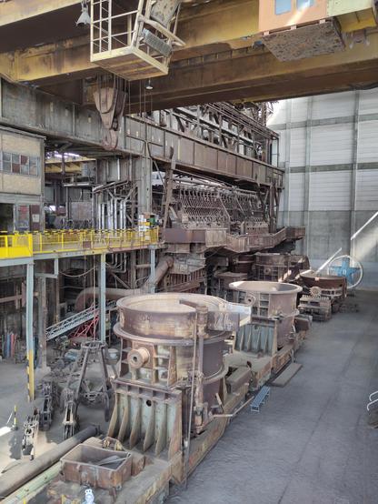 Das Bild zeigt das Innere eines ehemaligen Stahlwerks (das Industriemuseum Brandenburg), mit großen, rostigen Gießpfannen im Vordergrund, die auf Schienenwagen stehen und zum Transport von flüssigem Stahl dienten. Im Hintergrund erstreckt sich eine komplexe, alternde Hochofenanlage (der letzte erhaltene Siemens-Martin-Ofen)