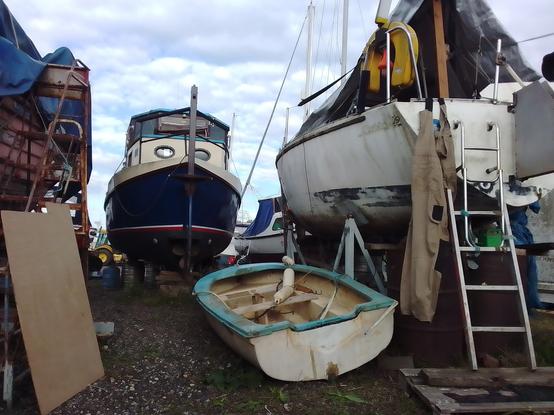A rowing dinghy beneath a tiny yacht in a cradle. A sheet of plywood to their left. Other boats in the background.