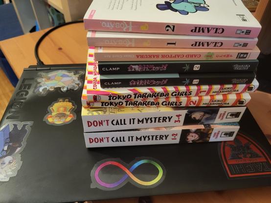 Ein Stapel Manga liegt auf einem mit Stickern verzierten schwarzen Laptop. Zu sehen sind Reihen wie Don't Call It Mystery, Tokyo Tarareba Girls, Legal Drug und Werke von CLAMP.
