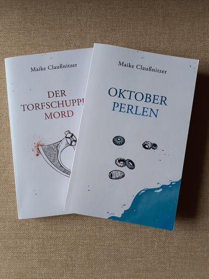 Auf beigefarbenem Hintergrund liegt das Buch "Oktoberperlen" schräg über dem Roman "Der Torfschuppenmord". Beide Cover sind von Sameena Jehanzeb (saje design) weißgrundig gestaltet. "Der Torfschuppenmord" hat als Covermotiv eine wikingerzeitlich anmutende Streitaxt, als Akzentfarbe dient Rot. "Oktoberperlen" hat eine Zeichnung von Glasperlen und Meerwasser, die Akzentfarbe ist Blau).