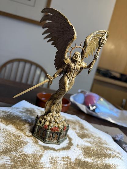 A piece of 28mm-scale statuary depicting a winged armoured woman holding a torch and sword, over a plinth topped with a pile of human skulls