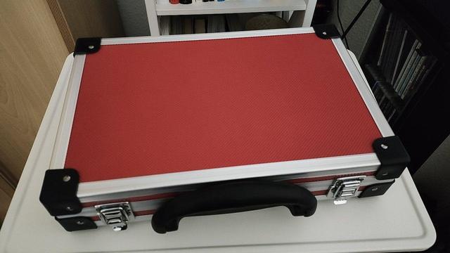 Ein kleiner flacher roter Gerätekoffer liegt geschlossen auf einem weißen Stehpult. Er ist nur ungefähr DIN A4 klein, deshalb wirkt der Griff überproportional groß.