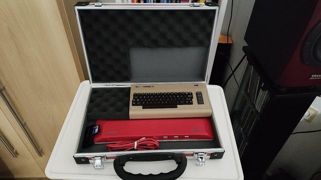 Der Deckel des Koffers ist  aufgeklappt. Im Boden sind zu sehen: 
1. Eine kleine Tastatur von Raspberry Pi. Sie liegt mit den Tasten nach unten, so dass der markante rote Boden nach oben schaut. 
2. Daneben ein zusammengelegtes USB-Kabel.
3. Daneben ein USB-Adapter mit einer SD-Karte.
4. Halb auf der Tastatur liegt der C64-Mini.
5. Daneben etwas Schaumstoff zum Fixieren.

Im Deckel ist Schaumstoff mit einer Aussparung passend zum C64-Mini eingelegt.