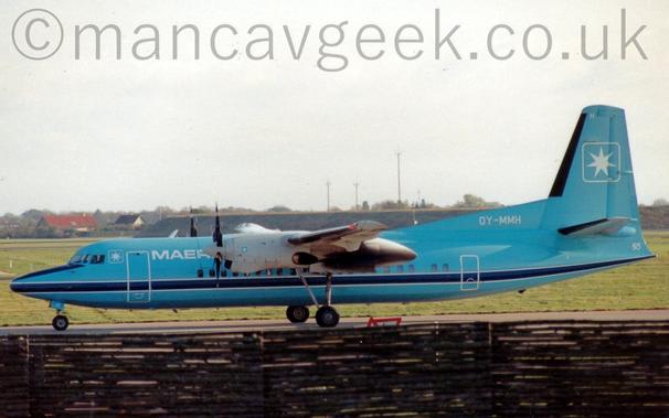 Side view of a light blue, high-winged, twin propellor-engined airliner taxiing from right to left.
The plane is mostly light blue, with a darker blue stripe outlined in white running along the body just below the passenger cabin windows.
There is the white outline of a square, with the corners rounded off, and containing a white, 7 pointed star, on the upper forward fuselage between the cockpit windows and the forward door, with white "Maersk Air" titles just aft of the door, although only the first 4 letters are visible, hidden behind the white engine pods poking out from the wings.
A larger version of the same square and star logo appears on the tail, with a smaller version on the side of the engine pods, just behind the propellors at the front.
The registration "OY-MMH" is on a fillet at the front of the tail, in white.
The main undercarriage is attached to open bays in the rear of the engine pods, on large retractable legs.
A brown wicker fence fills the foreground, the plane almost seeming to be taxiing along the top of it, with green grass in the background leading up to a grass covered berm in the background on the right, and red and brown roofs of houses on the left, under grey skies.