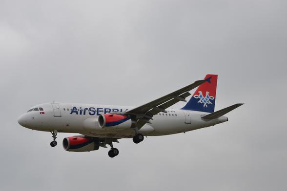 Side view of a white, twin engined jet airliner flying from right to left at a low altitude, with undercarriage lowered and flaps extended from behind the wings, suggesting it is about to land.
The plane has a white fuselage, with blue "Air Serbia" titles on the forward fuselage, covering some of the passenger cabin windows.
These titles are repeated in red under the fuselage, 
under the fairings where the wings meet the body.
The registration "YU-APF" is on the upper rear fuselage in black, repeated under the outer pane of the left wing.
The tail is red at the top and dark blue at the bottom, with an elaborate white outline of a 2 headed eagle with outstretched wings, the middle section coloured light blue.
The engine pods under the wings are red on top and white at the bottom, with a dark blue stripe swooping from the lower front to the upper rear.
Grey sky fills the rest of the frame.