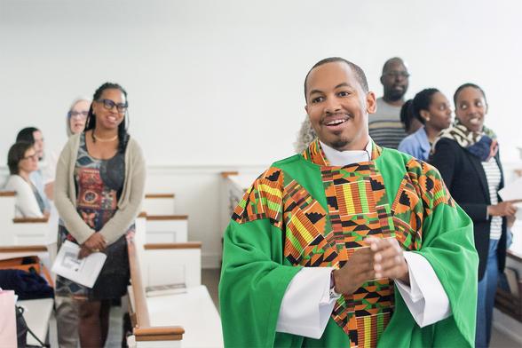A minister in brightly colored Kente cloth patterned liturgical robes, smiling, with congregants in the background.