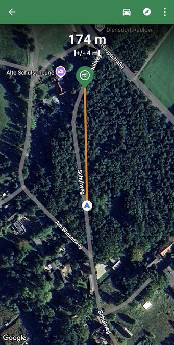 Satellitenbild aus einer Geocach Navigations-App. Eine orange Linie markiert einen 174 m langen Weg durch ein Waldstück auf dem „Schulweg“ in Diensdorf-Radlow. Ziel ist ein markierter Punkt bei der „Alten Schulscheune“. Die umliegenden Straßen und Häuser sind von dichtem Wald und Feldern umgeben.