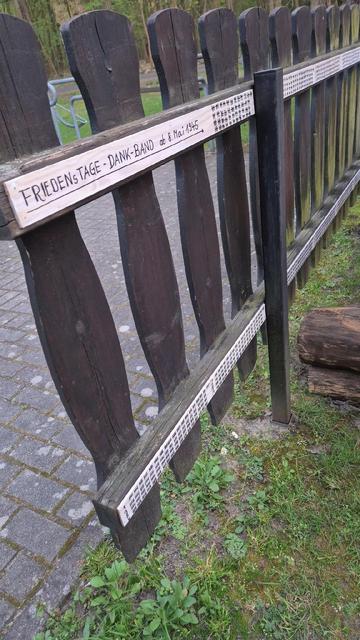 Ein dunkler Holzlattenzaun steht am Rand eines gepflasterten Wegs. Entlang einer der Zaunlatten ist ein helles Holzbrett befestigt, auf dem handschriftlich steht: „FRIEDENSTAGE – DANK-BAND ab 8. Mai 1945“. Darunter befinden sich hunderte kleine senkrechte Striche, die in regelmäßigen Abständen aufgemalt sind. Jeder Strich symbolisiert einen Tag in Frieden seit dem Ende des Zweiten Weltkriegs. Die Darstellung erstreckt sich entlang des gesamten Zaunfelds und macht die Friedenszeit seit 1945 sichtbar. Im Hintergrund sind Bäume und ein Park zu erkennen.