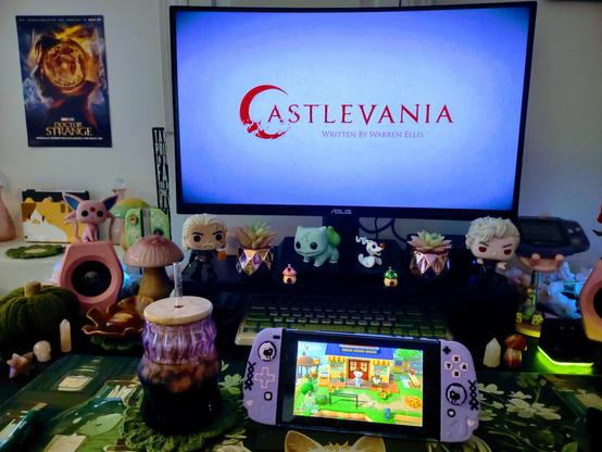 This is a photo of my gaming desk with sage and darker green shades, along with pops of pink. I have my Nintendo Switch with Animal Crossing's on the screen. It's sitting on my desk. I have Bulbasaur, Geralt, and Astarion Funko Pops beside my monitor, mushrooms everywhere, and crystals all around. Netflix's Castlevania is on the screen.