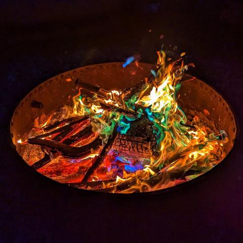 A close-up, top-down shot shows a vibrant fire burning within a rusty, oval-shaped fire pit against a dark background. The flames are a mesmerising mix of colours, including bright orange, yellow, red, blue, and green, swirling and dancing around burning logs and embers. Wisps of white smoke rise from the heart of the fire. The inside of the fire pit glows with the heat, contrasting sharply with the surrounding darkness. The colourful flames suggest the use of colour-enhancing fire packets, creating a captivating and almost magical effect.