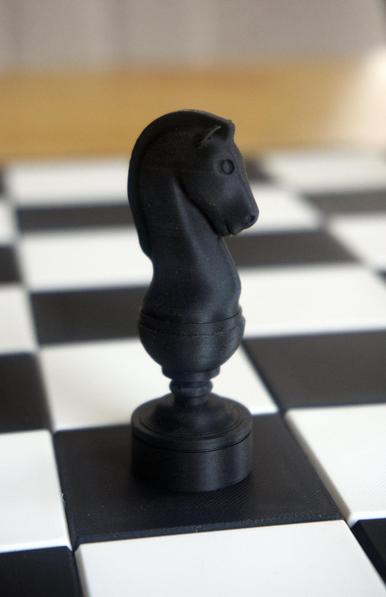 Closeup of a black Knight piece out of a 3D printed chess set and chess board. The photo has the knight in focus, the board is blurred.