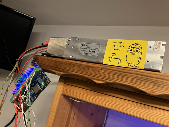 Foto eines Schaltnetzteils mit losen Kabel, welches an einer Platine angeschlossen ist. Auf dem Netzteil klebt ein gelber Warnsticker mit der Aufschrift: „Vorsicht da is Saft drauf!“