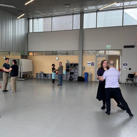 Three couples practice dancing, holding each other by the arms, while the teacher looks on. The nearest couple have their legs extended to the right.