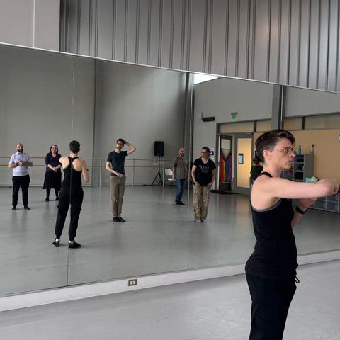 A short-haired person in a tank top raises their right arm in demonstration. Five students are visible in the mirror. A progress pride flag can also be seen hanging on the door, in the mirror.