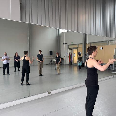 Six people look on, reflected in the mirror, as a person in a black tank top with short hair gesticulates, explaining.