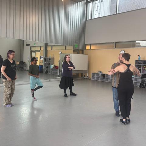 3 people watch as the teacher, dressed in a blank tank top and black pants, demonstrates a step with a student in a striped shirt.