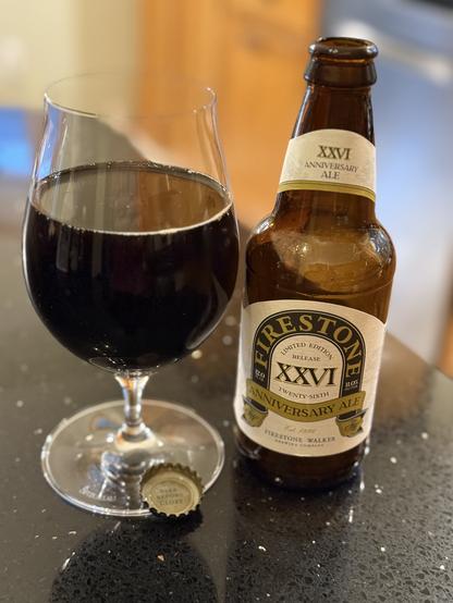 A snifter full of dark beer. The 12 ounce bottle is to the right with a white neck and body label. The Firestone logo is curved on a green field and the Roman numerals XXVI are n the middle