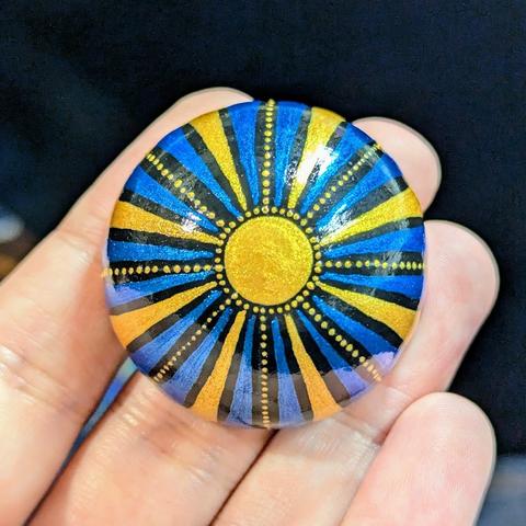 A small round half-sphere painted stone. The centre at the top is a gold circle, with radiating lines of gold dots, gold swooshes, and metallic blue swooshes arcing down to the bottom rim. It's glossed up and very shiny. Only about an inch and a half in diameter.