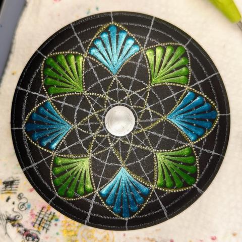An in progress dot painted stone. It's round and flat-ish, painted black, and covered in white and yellow guidelines drawn on with watercolour pencil. There's an eight-pointed geometric flower or star design made with small silver dots drawn on the grid, with the points of each "petal" filled with metallic swooshes in either green or teal. There's a large silver dot in the centre.