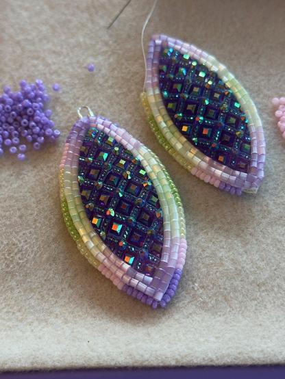 A pair of in progress beadwork earrings in purple, light pink, buttery yellow, and green, with dark purplish blue navette shaped centers.