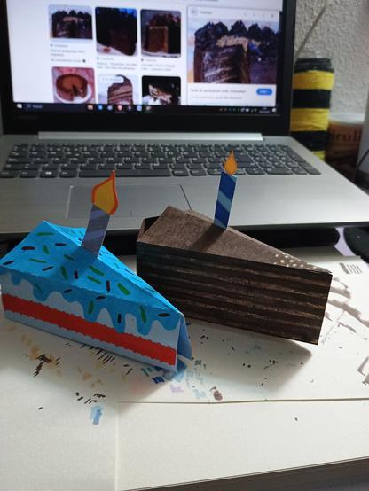 Fotografía de mi escritorio. En el se ven dos rebanadas de pastel en 3D que hice para una tarjeta de cumpleaños "pop up", hechas con cartulina de colores, cartulina blanca pintada y rotuladores.

A la izquierda está la "rebanada prototipo" que fue la primera que hice para ver como funcionaba y las medidas necesarias. A la derecha está la "rebanada final" que es la que pondré en la tarjeta de cumpleaños.

En el fondo se ve mi laptop con algunas imágenes de referencia que estoy utilizando para hacer la rebanada de pastel final.