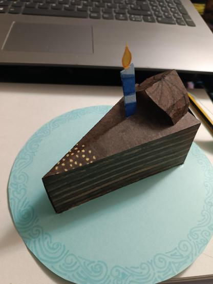 Fotografía de una rebanada de pastel de chocolate sobre un plato, el ambos hechos de cartulina. El pastel tiene una velita encendida, pedacitos de galleta y una decoración de chocolate en la parte superior

Son para una tarjeta pop up, así que se pueden doblar y quedar "planos", al abrirla se vería en 3D