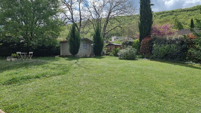 Un jardin avec sa pelouse taillée sur les trois quarts, une bande laissée en pousse libre , au fond un érable, des cyprès, des cabanes de jardin, à l'arrière plan le causse