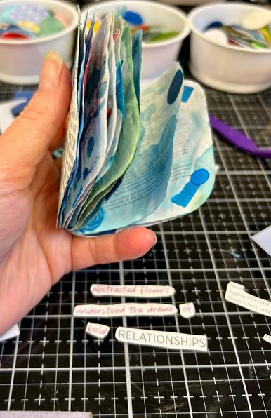 My pale hand holding a booklet of abstract collage. The pages are wavy from ink and glue on the very thin paper. A white on black gridded glass surface and recycled yogurt cups are in the background. There is a haiku of found text just below my hand on the glass.