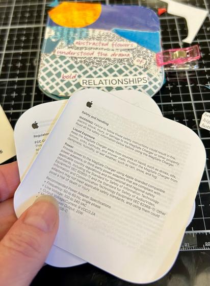 My hand holding product documentation from Apple. This is the type of booklet I covered with collage and found text haiku. The haiku booklet is in the background. I have placed a haiku on top of it, but it isn’t glued down yet. A pink clip is holding the haiku booklet closed.