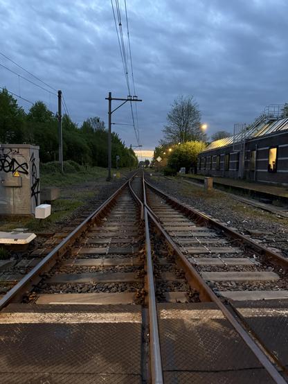 A perspective shot of some railroad tracks. The sky is moody. To the right, a building. To the left, some electrical boxes.