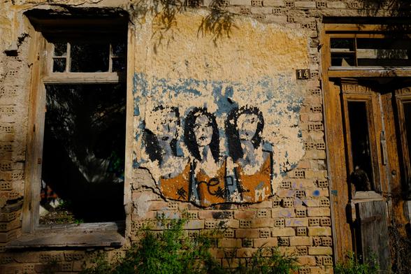 Photo of a decaying neo-classical house in golden sunlight. On its facade is a withered graffiti of three women.