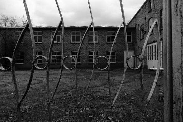 The image shows a black-and-white view of an old brick building with multiple windows. In the foreground, there's a decorative metal gate with curved bars and circular elements in its design. The sky looks cloudy.