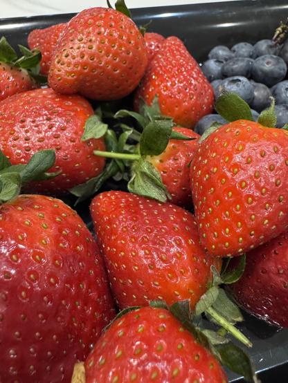 A small pile of juicy looking strawberries with their green stalks. There are some blueberries in the background