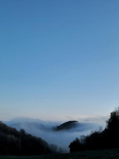 Sous un ciel bleu avant le lever du soleil, dans la perspective d'une vallée en V, le sommet d'une colline se détache de la brume qui "coule" autour.