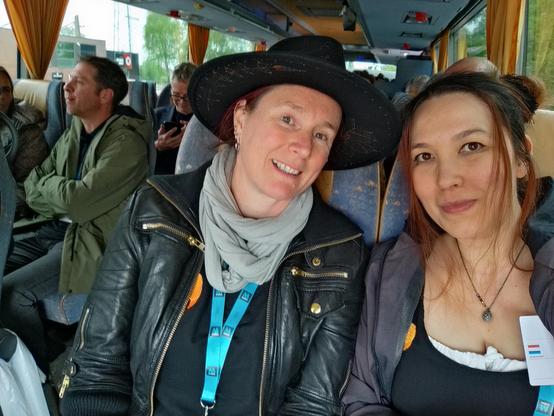 Two whitish women smiling in the front of a bus full of beer judges. The left woman wears a black hat, a light scarf, a leather jacket and the black polo from the last picture beneath, the right woman has her brown hair in two knot buns, a silver necklace with a hop bell hanger, a gray coat and the black dress underneath