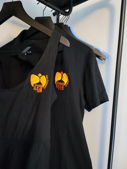 A black polo shirt with the original 10th anniversary logo for the Dutch Beer Challenge in yellow and orange embroidery and a black dress in front of it with a logo I embroidered myself on a piece of clothing I can wear without sensory issues