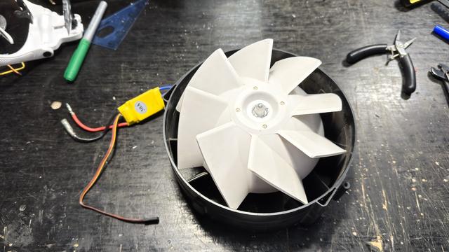 A jet-engine-looking turbine extractor fan that has been techromanced back to life from the innards of a burned out bathroom ceiling fan using a high-powered brushless drone motor and Electronic Speed Controller. The former is not visible in this picture, but the aerodynamic silver cap gives the white turbine a real spacey look. The latter, visible on the left, to one side of the tubular black housing, is a little yellow heatshrunk brick with red, blue, and black wires coming out of it. It sports a sticker that claims it can handle up to 30A of current delivery. At 12V, that's 360W of raw air-sucking power. This may be a very "Tim the toolman" undertaking... 😱 *obligatory grunting noises* #iykyk