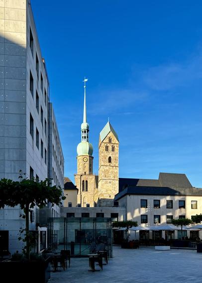 Ein moderner Platz mit einem Kontrast aus alter und neuer Architektur. Im Hintergrund erhebt sich die historische Reinoldikirche mit ihrem grünen Kupferdach, während im Vordergrund ein Gebäude mit glatter, dunkler Fassade steht. Der Platz ist mit Tischen, Stühlen und Bäumen gestaltet, die eine einladende Atmosphäre schaffen. Der klare blaue Himmel betont die Schönheit der Szene.