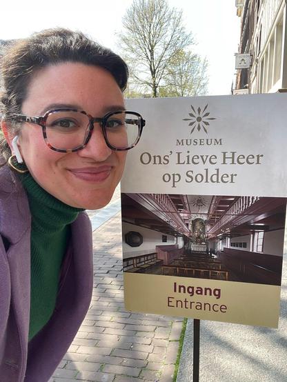 A smiling white woman with large glasses, earbuds, a green turtleneck, a purple jacket and dark hair in a bun. She looks glamorous and radiant. She's next to a sign for a museum written in Dutch.