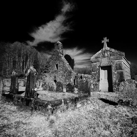 Black and white photo of the gable end and bell-cot of a ruined church. A large mausoleum sits next to it, topped with a cross shaped finial, behind tombstones.