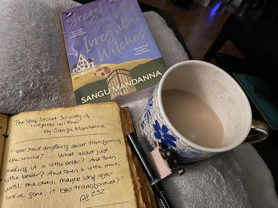 Three items sitting atop a lap covered in a white blanket.

1. A paperback copy of The Very Secret Society of Irregular Witches, by Sangu Mandanna (top center).
2. A cup of tea in a blue and gold floral mug (center right).
3. A journal with a quote from aforementioned book (bottom left). The quote reads as follows:

" 'Who said anything about transforming the world? ... What about just making it a little better? And then a little better? And then a little more, until, one day, maybe long after we're gone, it *has* transformed?' " –pg 232