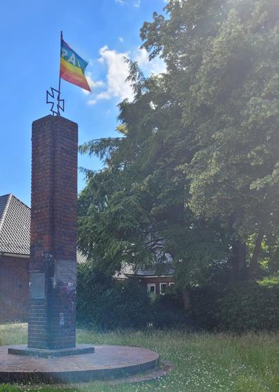 Auf einer kleinen, grünen, Gänseblümchenbewachsenen Wiese ist ein niedriger, gemauerter Kreis verlegt ,auf dem ein schmaler, eckiger Turm aus Klinker steht, ähnlich einem Schorstein. Oben auf dem Turm ist ein 'Eisernes Kreuz' angebracht, daran ist eine Ragenbogenfahne mit dem Schriftzug 'Pace' gehisst. Sie wwht im Wind. Im unteren Drittel des Turmes sind zwei Metalltafeln angebracht. Die eine ist unleserlich,  auf der anderen steht:"Den Toten aller Kriege". 
Im Hintergrund stehen große, sommergrüne Laubbäume und niedrige Häuser.