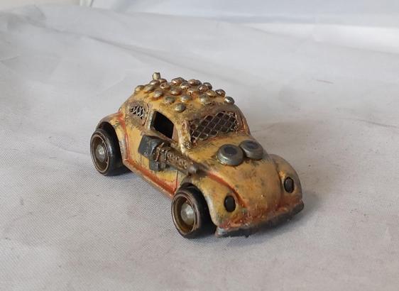 A Hot Wheels VW Beetle modified for the Gaslands game. Its mainly a rusty yellow, with a studded roof and a side-mounted machinegun. Front/angled view.