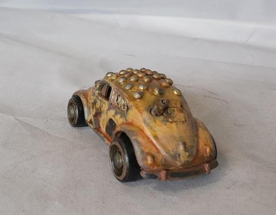 A Hot Wheels VW Beetle modified for the Gaslands game. Its mainly a rusty yellow, with a studded roof and a side-mounted machinegun. Rear/angled view.