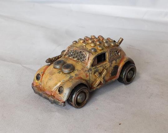 A Hot Wheels VW Beetle modified for the Gaslands game. Its mainly a rusty yellow, with a studded roof and a side-mounted machinegun. Front/angled view.