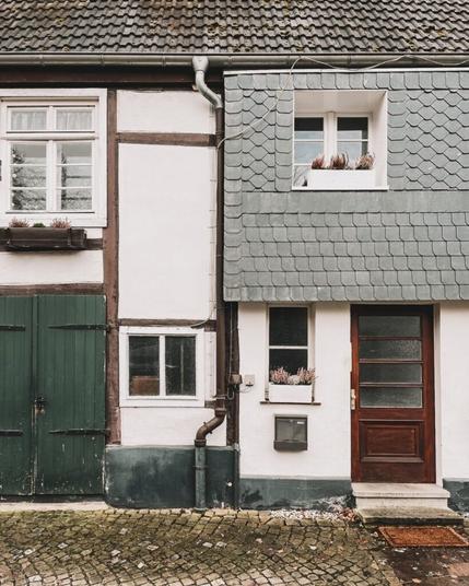 Die Fronten von zwei Fachwerkhäusern in Wiedenbrück. Man sieht jeweils nur die Hälfte. Weiße Fensterrahmen, eine grüne und eine braune Tür. In der Mitte sieht man ein Regenabwasserrohr. Auf den Fensterbänken des rechten Hauses stehen Blumen. Der Platz vor den Häusern ist aufwendig gepflastert.