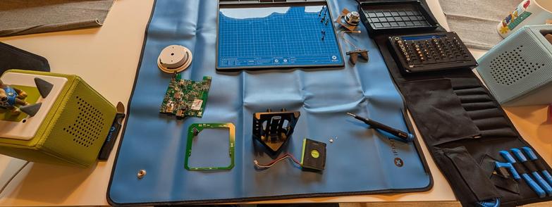 Zerlegte Toniebox auf einer blauen ESD Matte. Links daneben eine grüne Toniebox, rechts ein Werkzeugsatz von iFixit.