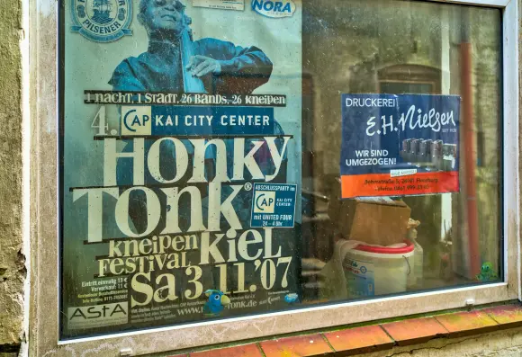 Ausschnitt aus einem Fenster in einem Hinterhof in Flensburg. Das Fenster ist dreckig, auf der Wand und dem Fensterbrett hat sich Moos angesammelt. Im Fenster hängt ein Plakat, das für das Honky Tonk Festival in Kiel 2007 wirbt, daneben ein Plakat, das anzeigt, dass die einst hier vorhandene Druckerei umgezogen ist.